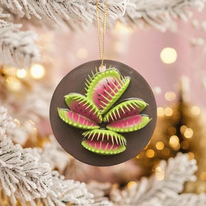 May include: A round, dark brown Christmas ornament featuring a stylized Venus flytrap design. The flytrap has bright pink and green coloring, with a gold string for hanging. The ornament is set against a blurred background of a Christmas tree.