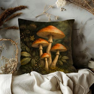 May include: Square throw pillow featuring a detailed illustration of several orange-capped mushrooms with beige stems, surrounded by green foliage. The pillow has a dark green background and is likely made of a soft fabric, perfect for home decor.