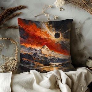 May include: A square throw pillow featuring a vibrant artistic print of a solar eclipse over a mountain range. The scene displays a dark moon surrounded by a bright sunburst against a fiery red and orange sky, with jagged mountains and a reflective lake.