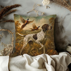 May include: A square throw pillow featuring a printed scene of a white and brown spotted hunting dog standing in a grassy field. Two pheasants are depicted in flight above the dog against a landscape background with trees and a cloudy sky.