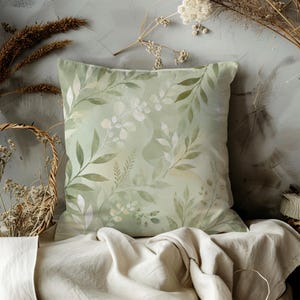 May include: Square decorative pillow with a watercolor botanical design. The pillow features a soft green background with a pattern of green and white leaves and branches. The pillow is placed on a cream-colored fabric.