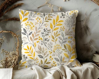 May include: Square throw pillow with a botanical pattern. The design features yellow, gray, and brown leaves and flowers on a cream background. The pillow is placed on a white fabric.