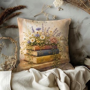 May include: A decorative throw pillow featuring a watercolor illustration of a stack of old books adorned with a floral arrangement. The flowers include daisies, lavender, and peonies in shades of white, purple, and pink. The pillow has a soft, neutral background.