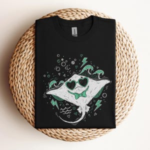May include: Black t-shirt featuring a cartoon stingray wearing heart-shaped sunglasses and a bow tie. The stingray is white with green accents, surrounded by bubbles, waves, and lightning bolts. The shirt is displayed on a woven, circular surface.
