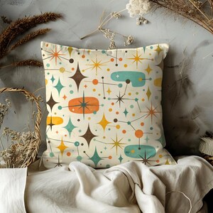 May include: Square decorative pillow with a retro atomic design. The off-white pillow features a repeating pattern of starbursts, dots, and oblong shapes in teal, orange, brown, and yellow. The pillow is placed on a white cloth.