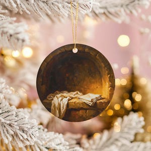 May include: Round Christmas ornament featuring a painting of a stone alcove with a draped cloth and folded cloth. The ornament has a gold string for hanging. The background is a blurred image of a Christmas tree.