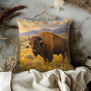 May include: Square throw pillow featuring a detailed painting of a bison in a grassy field with mountains in the background. The bison has brown fur and curved horns. The pillow has a neutral background.