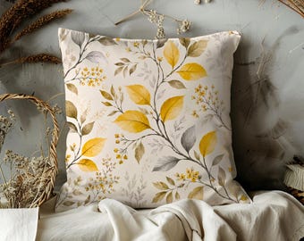 May include: Square decorative pillow with a botanical design. The pillow features a cream-colored background with a repeating pattern of yellow and gray leaves and small yellow flowers. The pillow is square and appears to be made of a soft fabric.