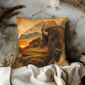 May include: A square throw pillow featuring a detailed print of a brown bison head in the foreground, set against a landscape of desert mesas, a winding river, and a golden sunset sky with distant mountains in warm orange and brown tones.