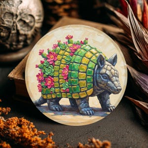 May include: A round coaster featuring an armadillo sculpture with a mosaic-like shell in green and yellow. The armadillo's back is adorned with pink flowers and green succulents. The coaster has a light beige background.
