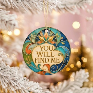 Peut inclure: Une décoration ronde et plate présentant une forme de cœur centrale de couleur crème avec le texte "YOU WILL FIND ME" en lettres sombres. Le motif est entouré de volutes complexes dans des tons de bleu sarcelle, bleu, orange et or.