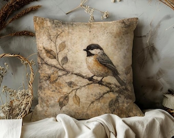May include: Square throw pillow with a vintage-style bird illustration. The pillow features a detailed painting of a small bird perched on a branch with leaves, set against a textured, neutral background. The color palette includes browns, tans, and muted greens.