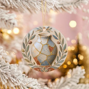 May include: A round ceramic ornament featuring a relief design of a blue and tan vase with gold-colored cracks. The vase is framed by a sculpted wreath of green olive branches and small brown olives, suspended by a thin gold-colored string.