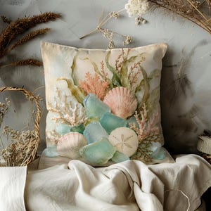 May include: A square throw cushion featuring a printed design of sea glass, seashells, a sand dollar, and coral branches in shades of light blue, peach, and cream. The imagery is arranged in a cluster against a soft, neutral-toned background.