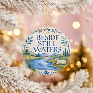 May include: A round ceramic ornament featuring a blue and white painted landscape with a river, rolling hills, and a sheep. The text BESIDE STILL WATERS is printed in blue in the center. The ornament hangs from a thin gold string.