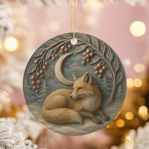 May include: A round Christmas ornament featuring a detailed illustration of a fox resting beneath a crescent moon and berry branches. The ornament has a blue-gray background with a gold hanging string.