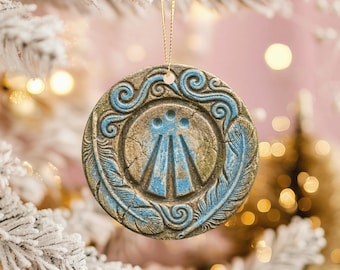 May include: A round, textured ceramic ornament featuring a central triskelion-inspired symbol in blue and weathered stone tones. The design includes embossed swirling patterns and two feather motifs along the edges, suspended by a thin gold cord.