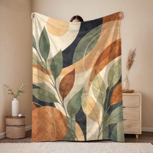 May include: A large rectangular throw blanket featuring an abstract botanical print with overlapping leaves in shades of sage green and terracotta orange. The design includes curved geometric shapes in cream, dark navy, and tan, creating a layered effect.
