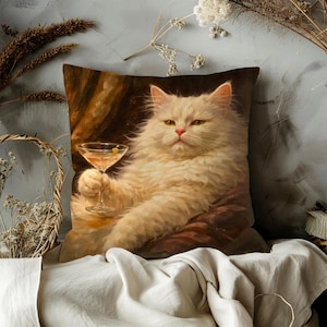 May include: Square decorative pillow featuring a regal white cat holding a cocktail. The cat has green eyes and a serious expression. The pillow has a brown and gold background, and the cat is the focal point.