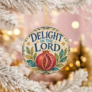 Peut inclure: Une décoration ronde en céramique avec le texte bleu "DELIGHT IN THE LORD" au-dessus d'une illustration centrale d'une grenade rouge coupée en deux avec des feuilles vertes. Le motif est bordé de décorations bleues et dorées et suspendu par un cordon doré.
