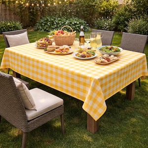 Puede incluir: Una mesa de picnic preparada para una reunión al aire libre, cubierta con un mantel a cuadros amarillos y blancos. La mesa está llena de comida, bebidas y una cesta de frutas. Sillas de mimbre rodean la mesa, sobre un césped.