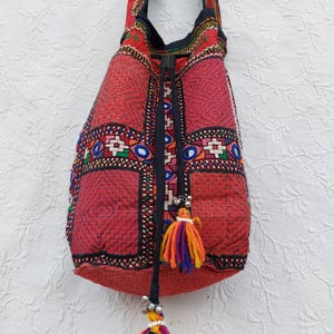 Könnte beinhalten: Eine rote Beuteltasche mit aufwendigen Stickereien in geometrischen Mustern, kleinen runden Spiegeln und einem Kordelzugverschluss. Die Tasche verfügt über einen langen Schulterriemen und zwei dekorative Quasten aus mehrfarbigem Garn.
