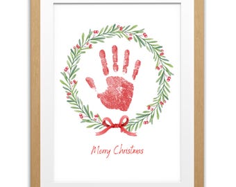 Puede incluir: Impresión enmarcada con una huella de mano roja dentro de una corona verde y roja. Una cinta roja está debajo de la corona, y las palabras "Merry Christmas" están escritas en rojo. La impresión está en un marco de madera marrón claro.