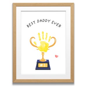 Pode incluir: Uma impressão emoldurada branca com o texto "Best Daddy Ever" e uma impressão de mão amarela dentro de um troféu dourado com uma base azul que diz "#1". Um pequeno coração rosa está no canto superior direito.