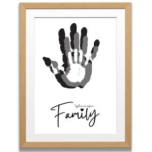 Può includere: Stampa incorniciata con un design di impronte di mani in tonalità di grigio e nero. La parola "Family" è scritta in corsivo sotto le impronte delle mani, con la frase "together we make a" sopra. La cornice è di colore marrone chiaro.