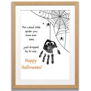 Pode incluir: Uma obra de arte emoldurada com tema de Halloween. A imagem apresenta uma impressão de mão transformada em uma aranha com olhos arregalados, pendurada em uma teia de aranha. O texto diz "The cutest little spider you have ever seen, just dropped by to say... Happy Halloween!"