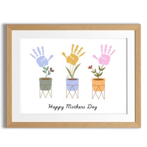 Puede incluir: Una impresión enmarcada con tres flores en macetas hechas con huellas de manos. Las huellas de manos son de color azul, amarillo y rosa. El texto "Happy Mothers Day" está en la parte inferior de la impresión.