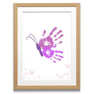 Può includere: Opera d'arte incorniciata con un disegno a farfalla realizzato con impronte di mani viola. Il corpo della farfalla è viola scuro, con antenne. Le ali sono formate da due serie di impronte di mani in tonalità di viola. Piccole sagome di farfalle sono sparse.