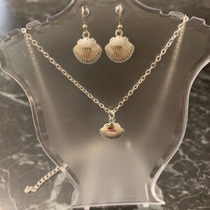 May include: A silver jewellery set comprising a seashell pendant necklace and matching seashell earrings. The necklace and earrings are displayed on a clear plastic stand against a dark background. The seashell pendants have a detailed, textured design.