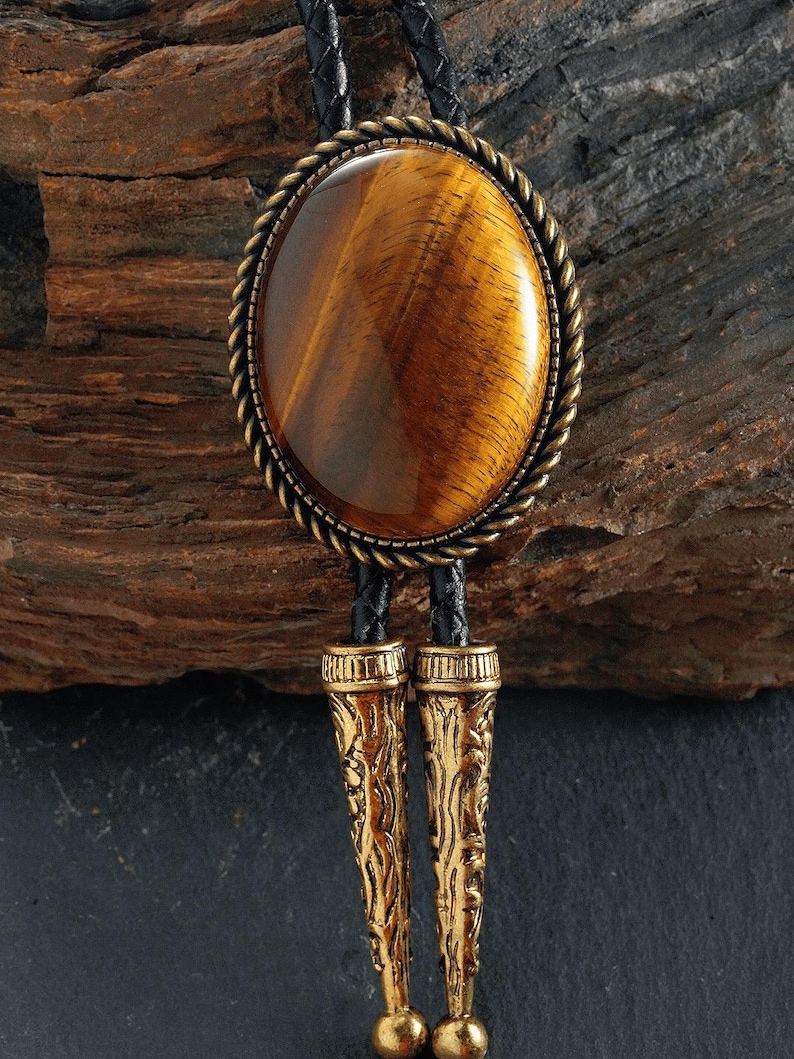 May include: A bolo tie with a large, oval tiger's eye stone set in a decorative, gold-toned metal frame with a rope-like border. The tie has a black leather cord and ornate gold-coloured metal tips.