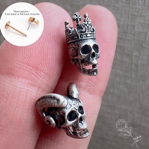 May include: Two silver skull-shaped earrings. One skull is crowned, the other has horns. The earrings are detailed with open mouths and eye sockets. A small inset shows push back or flat back closures.