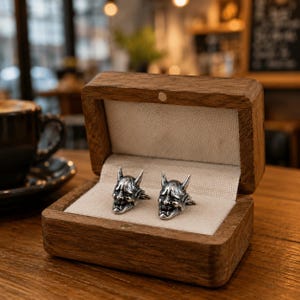 May include: A pair of silver-toned metal cufflinks shaped like Hannya masks, featuring intricate facial details and small horns. The cufflinks are displayed inside an open, rectangular wooden box with a light-coloured fabric lining.