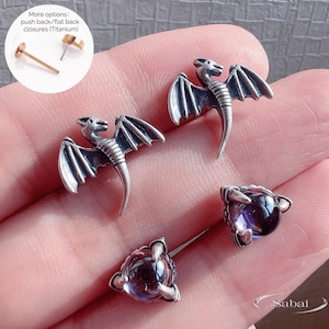 Pode incluir: Brincos de dragão prateados e brincos de botão de pedras preciosas roxas. Os brincos de dragão apresentam asas detalhadas e um corpo curvo. Os brincos de botão têm uma cravação em forma de garra. Opções de fecho adicionais são mostradas.