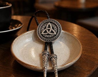 Könnte beinhalten: Eine Bolo-Krawatte mit einem kreisförmigen, silberfarbenen Anhänger, der ein zentrales Triquetra-Knotenmuster zeigt, umgeben von einem komplexen geflochtenen Rand. Der Anhänger ist an einer schwarzen geflochtenen Lederschnur befestigt, die mit zwei verzierten silberfarbenen Metallspitzen endet.