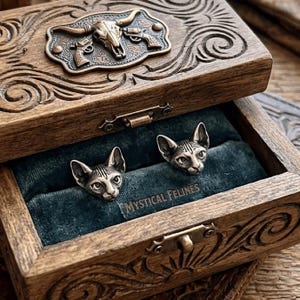 May include: A wooden jewelry box with an ornate carved design, featuring a bronze-colored metal emblem of a bull skull and pistols. Inside, two silver-colored cat head earrings rest on a teal velvet lining. The text "MYSTICAL FELINES" is visible.