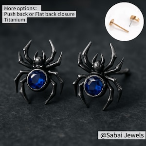 以下が含まれることがあります： 青い宝石のアクセントが付いた黒いクモのイヤリングのペア。イヤリングはチタン製で、プッシュバックまたはフラットバックの留め具が付いています。