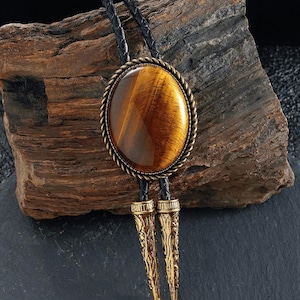 May include: A bolo tie with a large, oval tiger's eye stone set in a bronze-coloured metal frame with a rope design. The tie has a black leather cord and ornate, gold-coloured metal tips. The stone displays warm brown and gold tones.