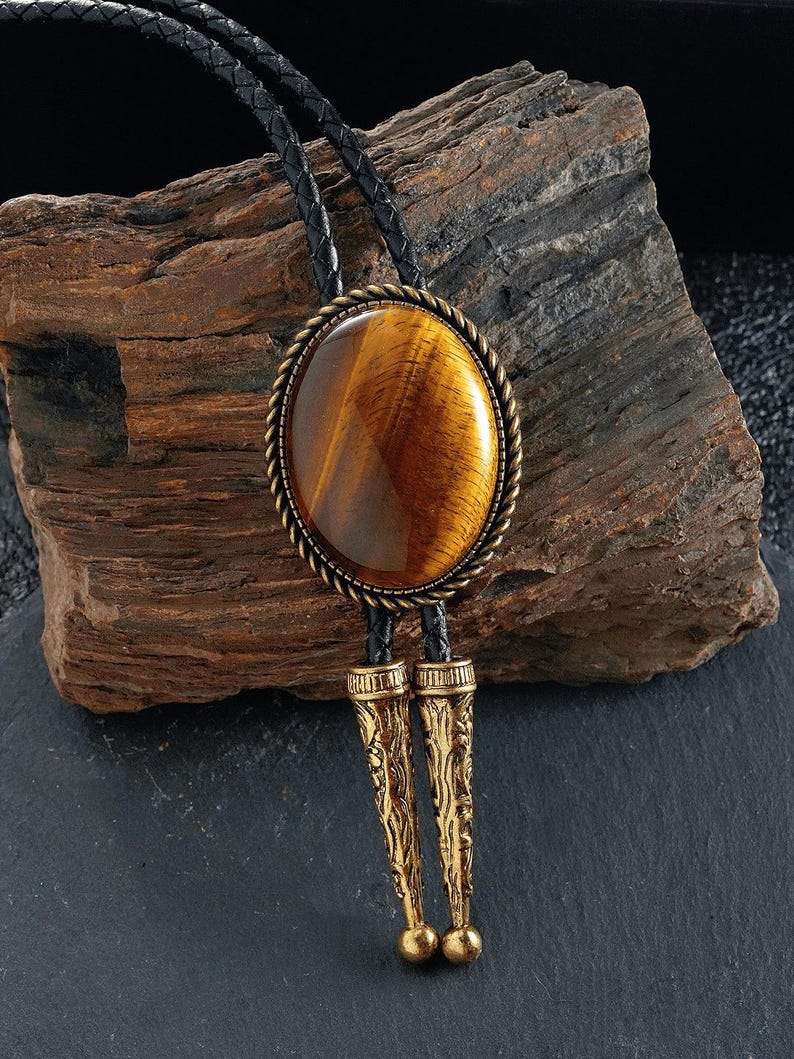 May include: A bolo tie with a large, oval tiger's eye stone set in a bronze-coloured metal frame with a rope design. The tie has a black leather cord and ornate, gold-coloured metal tips. The stone displays warm brown and gold tones.