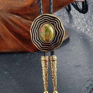 May include: A bolo tie featuring a central oval stone with green and red hues, set within a gold-toned, layered circular pendant. The pendant is suspended from a black braided leather cord, with decorative gold-toned tips.