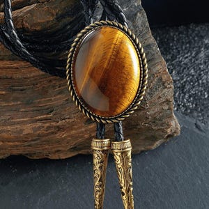 May include: A bolo tie with a large, oval tiger's eye stone set in a gold-toned metal frame with a rope-like border. The tie has a black braided leather cord and decorative gold-coloured metal tips. The stone has brown and gold hues.