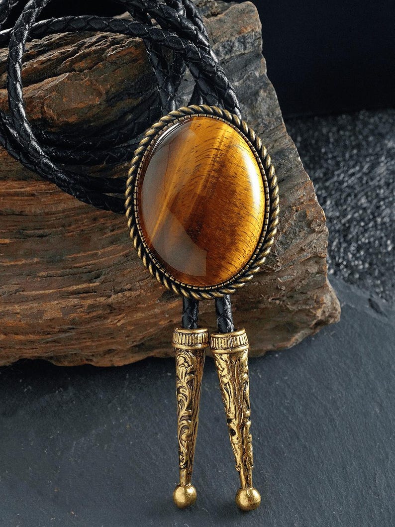 May include: A bolo tie with a large, oval tiger's eye stone set in a gold-toned metal frame with a rope-like border. The tie has a black braided leather cord and decorative gold-coloured metal tips. The stone has brown and gold hues.