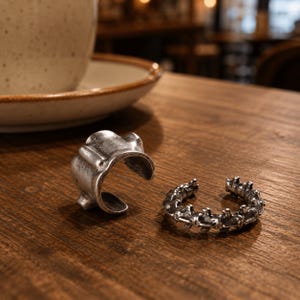 Pode incluir: Dois anéis de metal prateado com banda aberta. Um anel apresenta uma forma orgânica irregular e lisa com secções elevadas, enquanto o outro tem um design texturado e segmentado que se assemelha a uma série de pequenas vértebras conectadas.