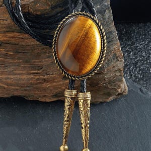 May include: A bolo tie featuring a large, oval tiger's eye stone set in a gold-toned metal frame with a rope-like border. The tie has a black braided leather cord and ornate gold-colored metal tips.