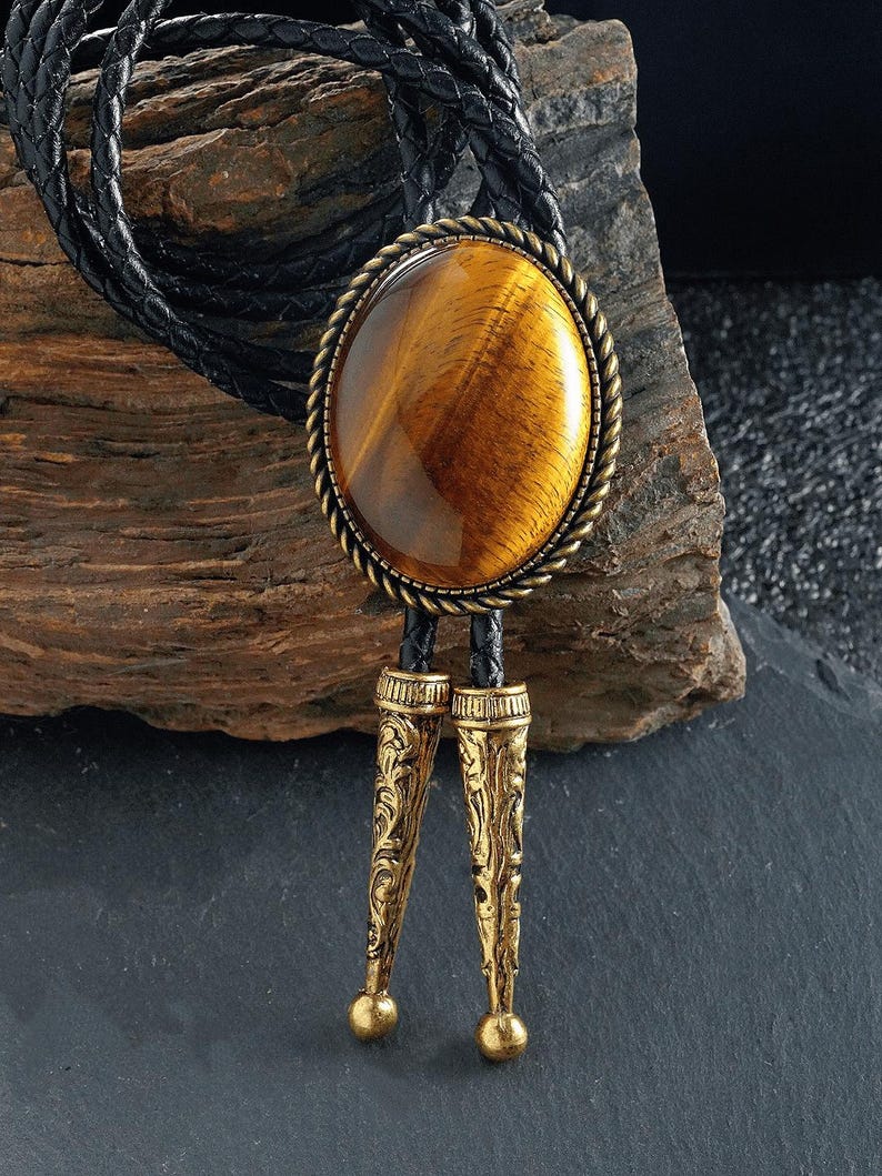 May include: A bolo tie with a large, oval tiger's eye stone set in a gold-toned metal frame with a rope-like border. The tie has a black braided leather cord and ornate gold-coloured metal tips.