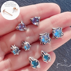 以下が含まれることがあります： イリデッセントブルーとパープルの宝石が付いた、シルバーカラーのスタッドピアスコレクション。ピアスは、三角形、正方形、有機的な形など、さまざまなデザインが特徴です。画像には、ゴールドトーンのプッシュバックピアスとテキスト「More options: push back/flat back closures (Titanium)」も表示されています。