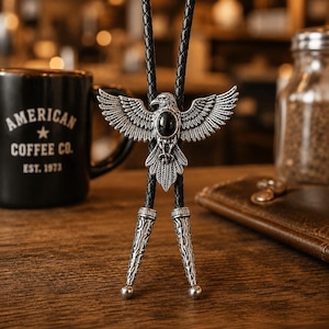 May include: A bolo tie featuring a silver-toned eagle pendant with an oval black stone at its center. The eagle has outstretched wings and detailed feather textures. The black braided cord ends in two matching silver-toned metal tips with engravings.